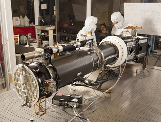 Photo of the instrument optics package prior to instrument level thermal vacuum testing.  The section to the left of the white collar is the 20cm solar telescope and the section to the right of the collar is the imaging spectrograph.  The spectrograph includes 18 optics used for transmitting the light from the telescope through the 4 channels to the focal planes as shown in the next sequence of images.  On top of the telescope assembly is the smaller guide telescope which provides the pointing signal to the secondary mirror of the telescope and to the attitude control system in the spacecraft.  The white collar is the primary mirror radiator used to reject the solar thermal load. Credit: LM Photo