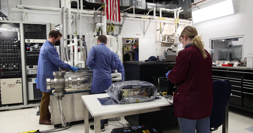 This video shows NASA engineers securing the BurstCube satellite in a thermal vacuum chamber for testing. The first shot shows a thermal vacuum chamber lab at NASA’s Goddard Space Flight Center in Greenbelt, Maryland. The second clip shows Julie Cox (NASA) and Seth Abramczyk (NASA) moving BurstCube, still in a clear protective case, from a table to the chamber platform. The next shot shows Cox and Abramczyk talking over the spacecraft, now without the covering. NASA engineers Franklin Robinson and Elliot Schwartz look on. In the fourth and fifth shots, all four engineers work to move BurstCube into position. In the sixth shot, Cox props one of the solar panels slightly open so they can test it when the spacecraft is in the chamber. In the next shot, Cox, Abramczyk, and Robinson make more adjustments. The eighth shot shows one side of BurstCube, which is engraved with the mission’s logo and the names of partner institutions. The following two clips show wider views of the spacecraft on the chamber platform. The eleventh shot shows Abramczyk and Cox typing at their computers. The twelfth shot shows a computer screen with a feed from a camera on the chamber platform. A smiling Abramczyk pops in and out of view. The thirteenth clip shows Cox deploying one solar panel with Abramczyk and Robinson in the background. The fourteenth shot shows Schwartz, Robinson, Abramczyk, and Colton Cohill (NASA) moving the top of the chamber into place. The fifteenth through nineteenth shots show the engineers steadying the lid as it lowers slowly into place. Shot twenty shows Schwartz securing the top of the lid. Shot twenty-one shows a pan of the sealed chamber. The final shot shows a data readout on a computer screen.
Credit: NASA/Sophia Roberts