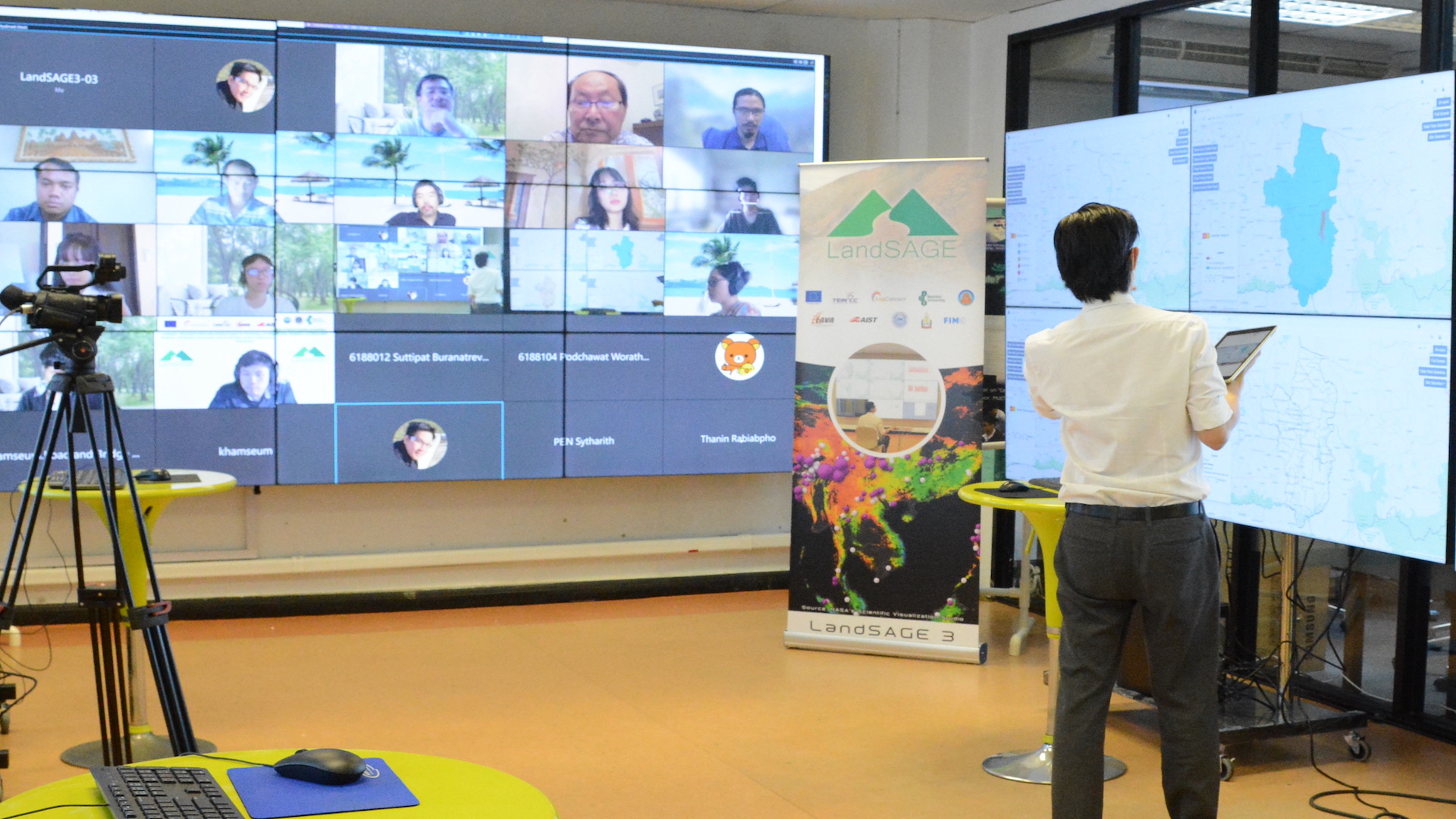 A man is giving a presentation in front of a screen. Visible in the background is a number of people teleconferencing in