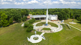 An afternoon view above the rocket garden at NASA Goddard's Visitor Center. The prominent Delta B launch vehicle stands 90-feet tall and was a type used to orbit satellites in the early 1960s. In the foreground, a kinetic sculpture called Orbits Interweave incorporates three polished stainless steel spheres &mdash; representing the Sun, Earth, and GOES weather satellites &mdash; that move gently in the wind. The exhibit area around it is shaped like a hurricane symbol. Imaged June 13, 2023, looking east-southeast.Credit: NASA/Francis Reddy